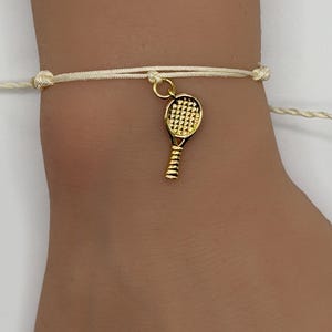 May include: A tennis-themed bracelet featuring a gold-colored tennis racket charm. The charm is suspended from a cream-colored cord bracelet. The bracelet is tied with a knot closure.