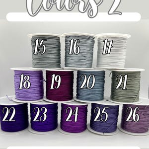 Puede incluir: Una selecci&oacute;n de carretes de cord&oacute;n de colores, cada uno numerado del 15 al 26. Los cordones son de varios tonos de gris, morado y verde. El texto "Colors 2" est&aacute; en la parte superior.