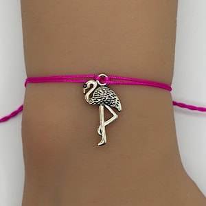 May include: A pink string bracelet with a silver flamingo charm. The flamingo charm has a detailed design and is attached to the bracelet with a small ring. The bracelet is worn on a wrist.