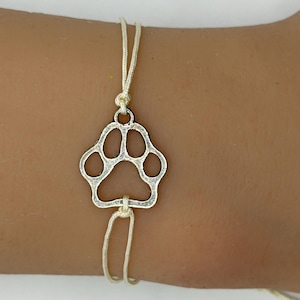 May include: Silver-colored paw print charm bracelet. The charm is a cutout of a paw print, suspended on a thin, adjustable, off-white cord. The bracelet is designed to be worn on the wrist.