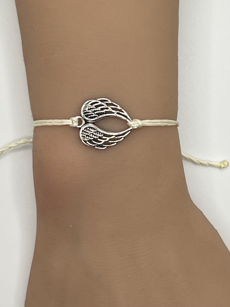 Puede incluir: Una pulsera con dije de color plateado con un par de alas de &aacute;ngel estilizadas. Las alas est&aacute;n conectadas a una pulsera de cord&oacute;n de color crema. La pulsera est&aacute; atada con un cierre de nudo corredizo.