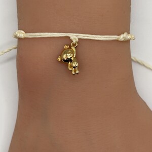 May include: A gold-colored bear charm bracelet. The charm is suspended from a cream-colored cord bracelet. The bracelet is tied with knots on each side. The bear charm is detailed with visible features.