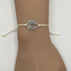 Puede incluir: Una pulsera con dije plateado con un par de alas de &aacute;ngel estilizadas. Las alas est&aacute;n conectadas a un cord&oacute;n de color crema, creando un dise&ntilde;o simple pero elegante. La pulsera se usa en una mu&ntilde;eca.