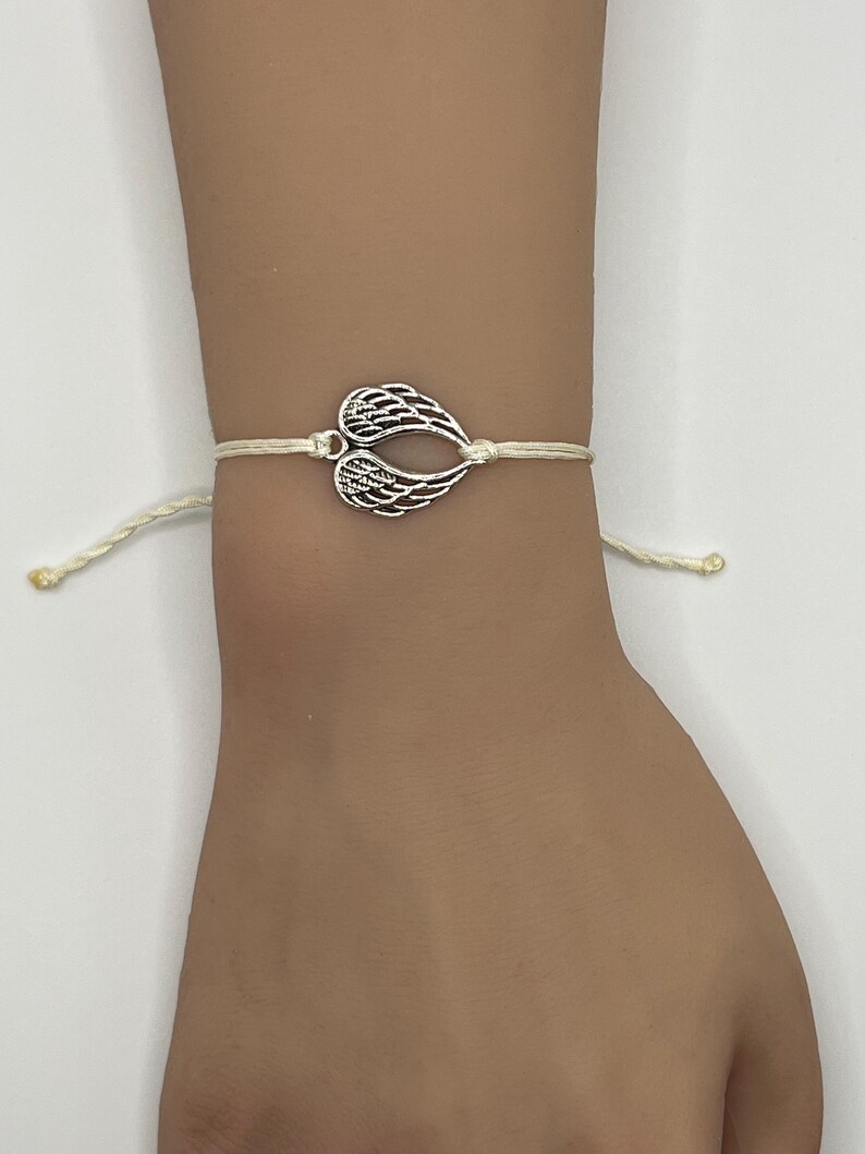 Puede incluir: Una pulsera con dije plateado con un par de alas de &aacute;ngel estilizadas. Las alas est&aacute;n conectadas a un cord&oacute;n de color crema, creando un dise&ntilde;o simple pero elegante. La pulsera se usa en una mu&ntilde;eca.