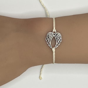 Puede incluir: Una pulsera con un dije de alas de &aacute;ngel plateadas. El dije est&aacute; centrado en un cord&oacute;n color crema. La pulsera est&aacute; atada con un nudo y tiene una peque&ntilde;a cuenta dorada al final del cord&oacute;n.