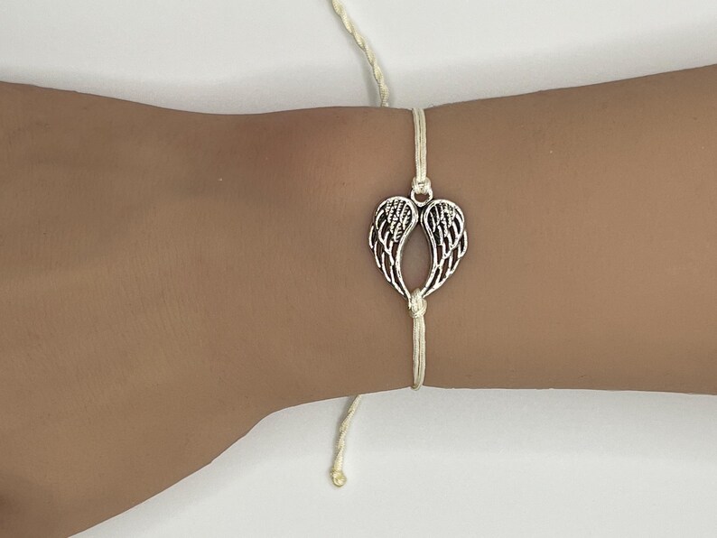 Puede incluir: Una pulsera con un dije de alas de &aacute;ngel plateadas. El dije est&aacute; centrado en un cord&oacute;n color crema. La pulsera est&aacute; atada con un nudo y tiene una peque&ntilde;a cuenta dorada al final del cord&oacute;n.