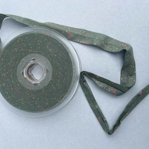 Könnte beinhalten: Eine Rolle olivgrünes Schrägband mit einem gesprenkelten Muster aus roten und gelben Punkten. Das Band ist abgewickelt und zeigt die Textur und Farbvariationen des Stoffes. Das Band befindet sich auf einer transparenten Kunststoffspule.