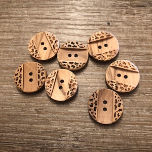 Könnte beinhalten: Sieben Holzknöpfe mit natürlicher Holzmaserung. Jeder Knopf hat zwei Löcher zum Annähen und ein dekoratives geätztes Design. Die Designs umfassen einen Leopardenmuster und vertikale Streifen. Die Knöpfe sind auf einer Holzoberfläche angeordnet.