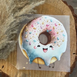 Könnte beinhalten: Ein Cartoon-Donut mit weißem Zuckerguss, rosa und blauen Streuseln und einer Schokoladenmitte. Der Donut hat ein fröhliches Gesicht mit großen Augen und einem breiten Lächeln. Er hat zwei kleine Beine und steht auf einer hellbraunen Oberfläche.