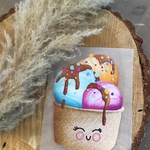 Könnte beinhalten: Eine skurrile Illustration einer Eistüte mit drei Kugeln. Die Kugeln sind blau, orange und lila, garniert mit Schokoladensauce und Streuseln. Der Kegel hat ein lächelndes Gesicht. Das Bild befindet sich auf einem Stück Papier.