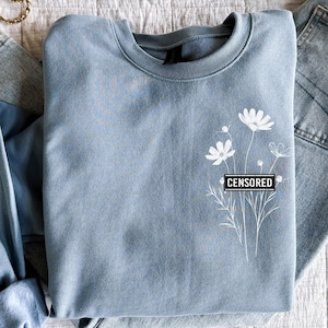 May include: A light blue sweatshirt with a white floral design and the word "CENSORED" printed in black on the front. The sweatshirt is folded and laid on a white textured surface with a pair of gold hoop earrings.