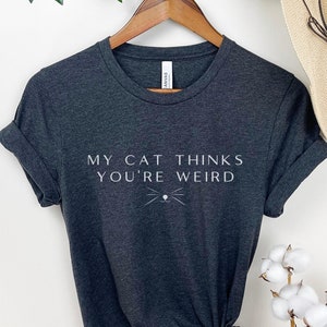 May include: A dark gray t-shirt with the text "My cat thinks you're weird" in white lettering. A stylized cat face with whiskers is below the text.