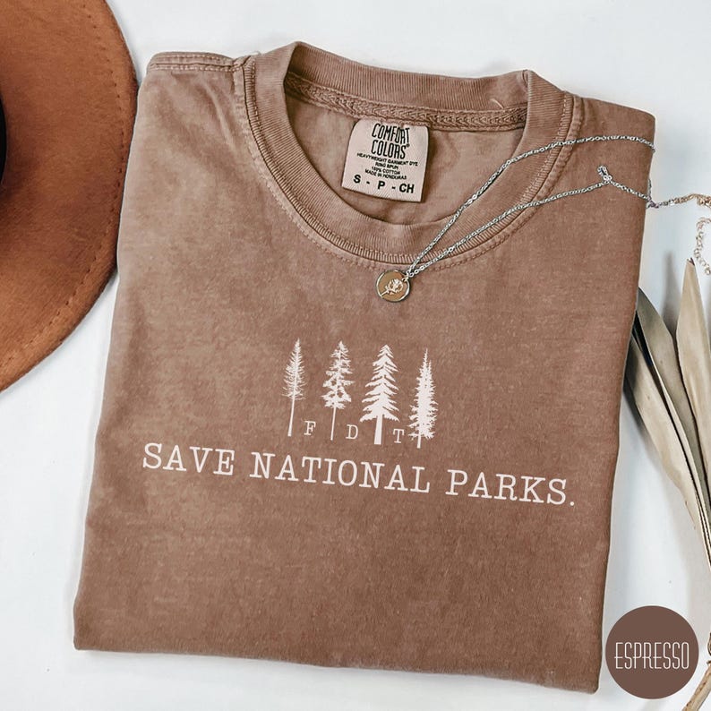 May include: A brown t-shirt with a white graphic of five trees and the text "SAVE NATIONAL PARKS." The shirt also has a white tag with the text "COMFORT COLORS" and "S - P - CH".