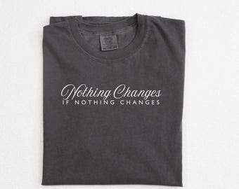 May include: A dark gray t-shirt with the words "Nothing Changes IF NOTHING CHANGES" in white script. The shirt is folded neatly, and the color is labeled "Pepper" at the bottom right.