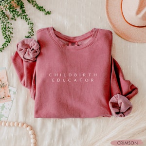 May include: Crimson-colored sweatshirt with the text "CHILDBIRTH EDUCATOR" in white. The sweatshirt is folded, with the sleeves and neckline visible. A scrunchie in the same color is next to the sweatshirt. A hat and greenery are in the background.