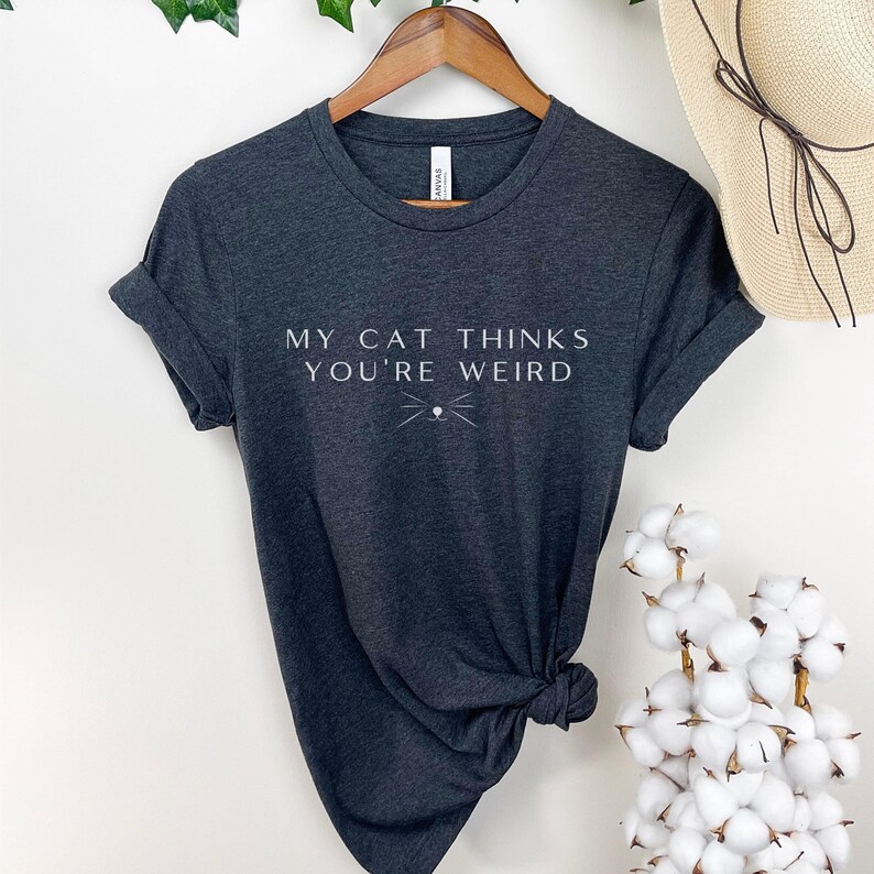 May include: A dark gray t-shirt with the text "My cat thinks you're weird" in white lettering. A stylized cat face with whiskers is below the text.