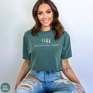 May include: A woman wearing a dark green t-shirt with the text "Save National Parks." and a line drawing of trees. She is wearing blue jeans with distressed details.