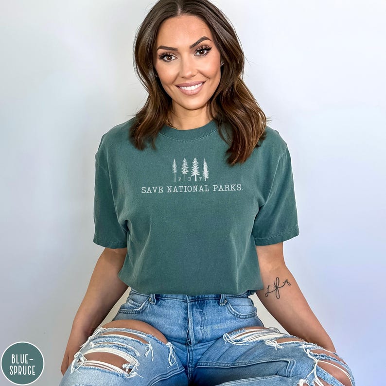 May include: A woman wearing a dark green t-shirt with the text "Save National Parks." and a line drawing of trees. She is wearing blue jeans with distressed details.