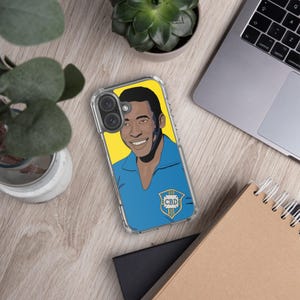 May include: Clear phone case featuring a smiling portrait of a man in a blue shirt against a yellow background. The case has a blue and green emblem with the letters "CBD". The case is on a wooden surface with a laptop and plants.