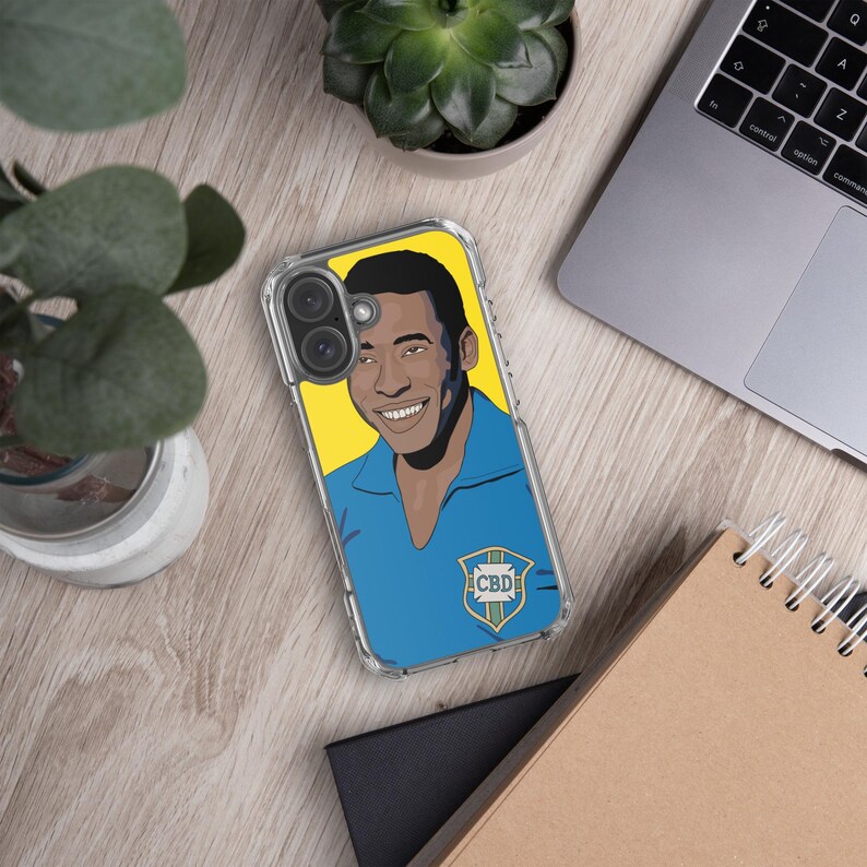 May include: Clear phone case featuring a smiling portrait of a man in a blue shirt against a yellow background. The case has a blue and green emblem with the letters "CBD". The case is on a wooden surface with a laptop and plants.