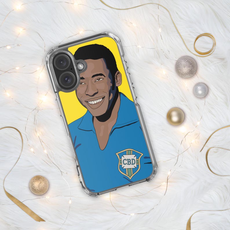 May include: Clear phone case featuring a portrait of Pel&eacute; in a blue jersey with a yellow background. The case has a clear bumper and a graphic of the CBD logo. The phone case is on a white fluffy surface with gold and silver decorative elements.