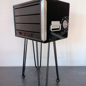 May include: A black, glossy cabinet with four drawers and a circular porthole detail. The cabinet has a metal handle and is supported by black hairpin legs. The edges are reinforced with silver metal. A unique piece of furniture.