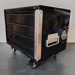 May include: A black metal rolling tool chest with three drawers, a side handle, and a circular access port. The chest has a shiny finish and is accented with silver metal hinges and trim. It sits on four small caster wheels.