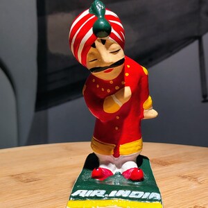 May include: A painted figurine of a person wearing a red and white striped turban and a red tunic with yellow trim. The figure has a black mustache, closed eyes, and is bowing. It stands on a green base with the white text AIR INDIA printed on it.