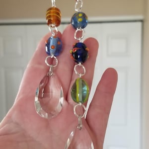 May include: Two decorative beaded crystal suncatchers. Each suncatcher features a teardrop-shaped crystal pendant and a strand of colorful glass beads in shades of brown, blue, green, and orange. The beads are connected by silver-colored metal rings.