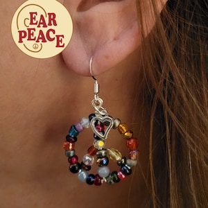 May include: Peace sign earrings made with colorful beads and a silver heart charm. The earrings are hanging from a person's ear.