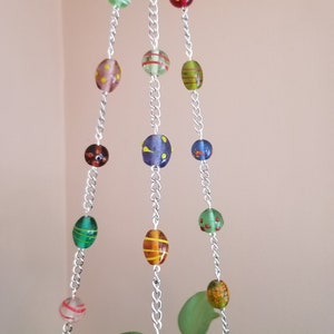 May include: Three strands of silver chain with colorful glass beads hanging from the ceiling. The beads are various shades of green, red, yellow, and blue with different patterns. The chains are hanging over a green leafy plant.
