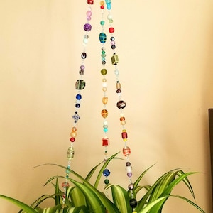 May include: A hanging plant holder made of colorful glass beads. The holder is attached to a ceiling hook and is hanging over a green plant with white stripes.