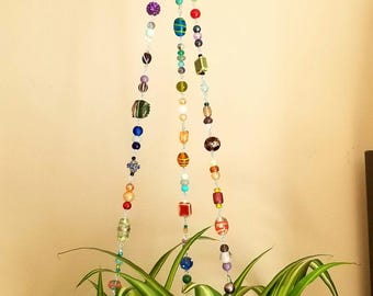 May include: A hanging plant holder made of colorful glass beads. The holder is attached to a ceiling hook and is hanging over a green plant with white stripes.