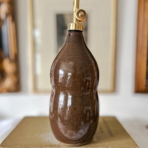 May include: A brown ceramic oil cruet with a glossy finish and a bulbous, rounded body shape. The bottle is topped with a metallic gold-colored pourer spout featuring a small circular hinge mechanism at the top for controlled liquid dispensing.