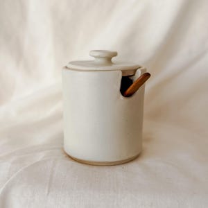 May include: A cylindrical off-white ceramic jar with a matching lid and a small notch on the rim. A wooden spoon is inserted into the jar through the notch, with the handle protruding outward. The jar has a smooth, matte finish and a flat base.