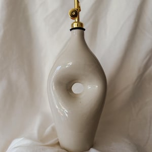 Peut inclure: Une burette à huile en céramique blanc cassé brillant avec une découpe circulaire centrale distinctive. Le récipient présente un col effilé surmonté d'un bec verseur doré métallique, incluant un petit mécanisme à charnière circulaire.