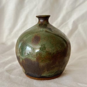 May include: A small, round ceramic vase with a narrow neck and a flared rim. The surface features a mottled glaze with blended tones of moss green, dark brown, and black, revealing a smooth, glossy finish over a textured, unglazed clay base.