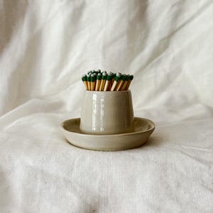 May include: A beige ceramic match holder with a matching circular saucer base, containing a cluster of wooden matches with green tips. The vessel has a smooth, glossy finish and a tapered cylindrical shape, holding the matches upright.