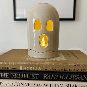 May include: A ceramic ghost-shaped candle holder with three cut-out openings for eyes and mouth, and a small candle inside. The candle holder is beige and sits on a stack of books with gold lettering. The books are titled "Conversational Russian", "The Prophet", and "Sonnets".
