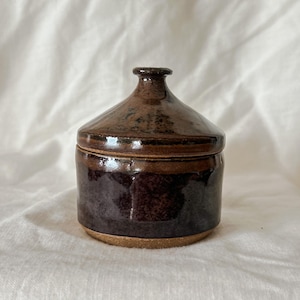 May include: A small, cylindrical ceramic jar with a matching lid, featuring a glossy, dark brown glaze with subtle black speckling. The base is unglazed, revealing a natural tan clay texture, and the lid has a small, centred knob handle.