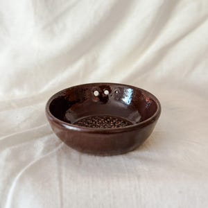 May include: A small, round, dark brown ceramic bowl with a glossy finish. The interior base features a textured, grater-like surface for processing ingredients. Two small, circular holes are positioned near the upper rim of the bowl.