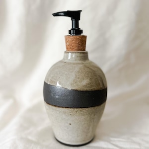 May include: A ceramic soap dispenser with a black pump and a cork stopper. The dispenser has a light gray base with a dark gray band around the middle. The dispenser is a decorative bathroom accessory.