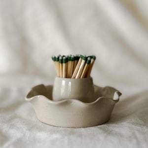 May include: A ceramic match holder and tray set. The set is a light beige color. The match holder is a small, cylindrical vessel, and the tray has a scalloped edge. The match heads are green.