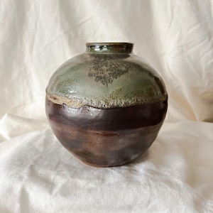 May include: A handcrafted ceramic vase with a rounded body and a narrow neck. The upper portion is a mottled green, transitioning to a dark brown base. The vase has a textured surface and a rustic aesthetic, suitable for home decor.
