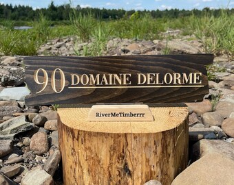 May include: A dark brown wooden sign with the text "90 Domaine Delorme" in gold lettering. The sign is sitting on a small wooden stand with the text "RiverMeTimberrr" in black lettering. The sign is sitting on a pile of rocks.