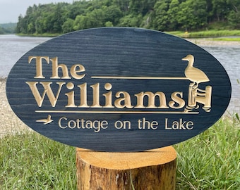 May include: A dark blue oval wooden sign with the text "The Williams Cottage on the Lake" carved into it. A small bird silhouette is carved above the text. The sign is mounted on a natural wood log.