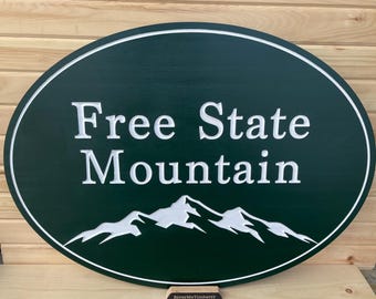May include: Oval-shaped wooden sign with a green background and white lettering. The sign reads "Free State Mountain" with a stylized mountain range graphic below the text. The sign is on a wooden stand.