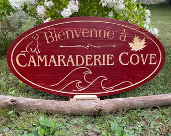 May include: A red wooden oval sign with the text "Bienvenue à CAMARADERIE COVE" in white lettering. The sign also features a dog silhouette, a maple leaf, and stylized waves.