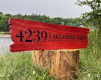 May include: A red wooden sign with black lettering that reads "4239 LAKESHORE DRIVE". The sign is sitting on a tree stump in a grassy area.