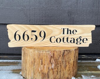 May include: A wooden address sign with the numbers "6659" and the words "The Cottage" in black lettering. The sign has a natural wood grain finish and is displayed on a tree stump. The background is a dark wooden wall.